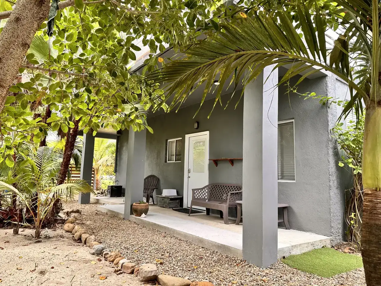 Studio porch shaded by palms, with chairs beside the grey house